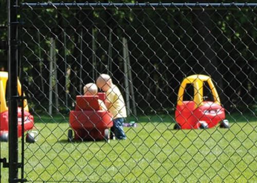 A child is playing with a toy car in a yard behind a chain link fence