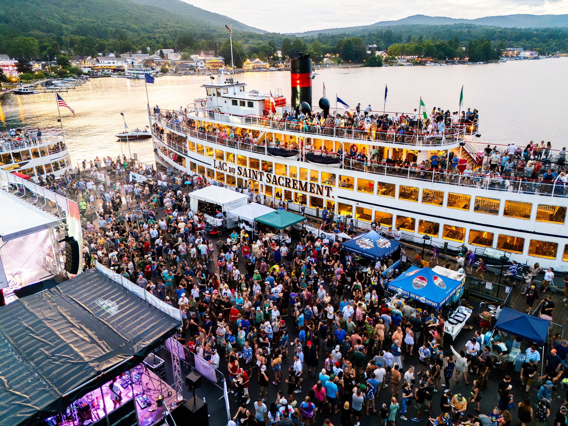 Crowd at outdoor music festival near a large boat. People raise hands, smiling. Some wear colorful clothing.