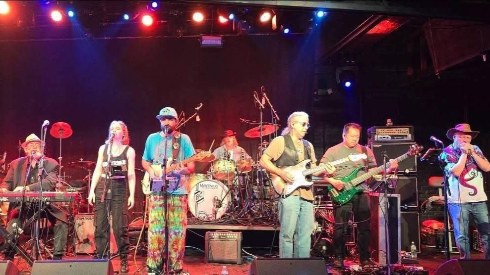 Band on stage, lit with colorful lights. Musicians play instruments: keyboard, drums, guitars, violin.