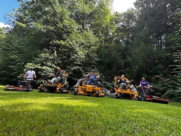 Five people on riding lawn mowers, parked on grass, with a backdrop of lush green trees under a sunny sky.