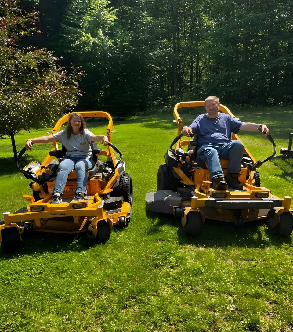 Two people Husband & Wife sit on yellow riding lawn mowers on a bright green lawn, surrounded by lush trees and bushes.