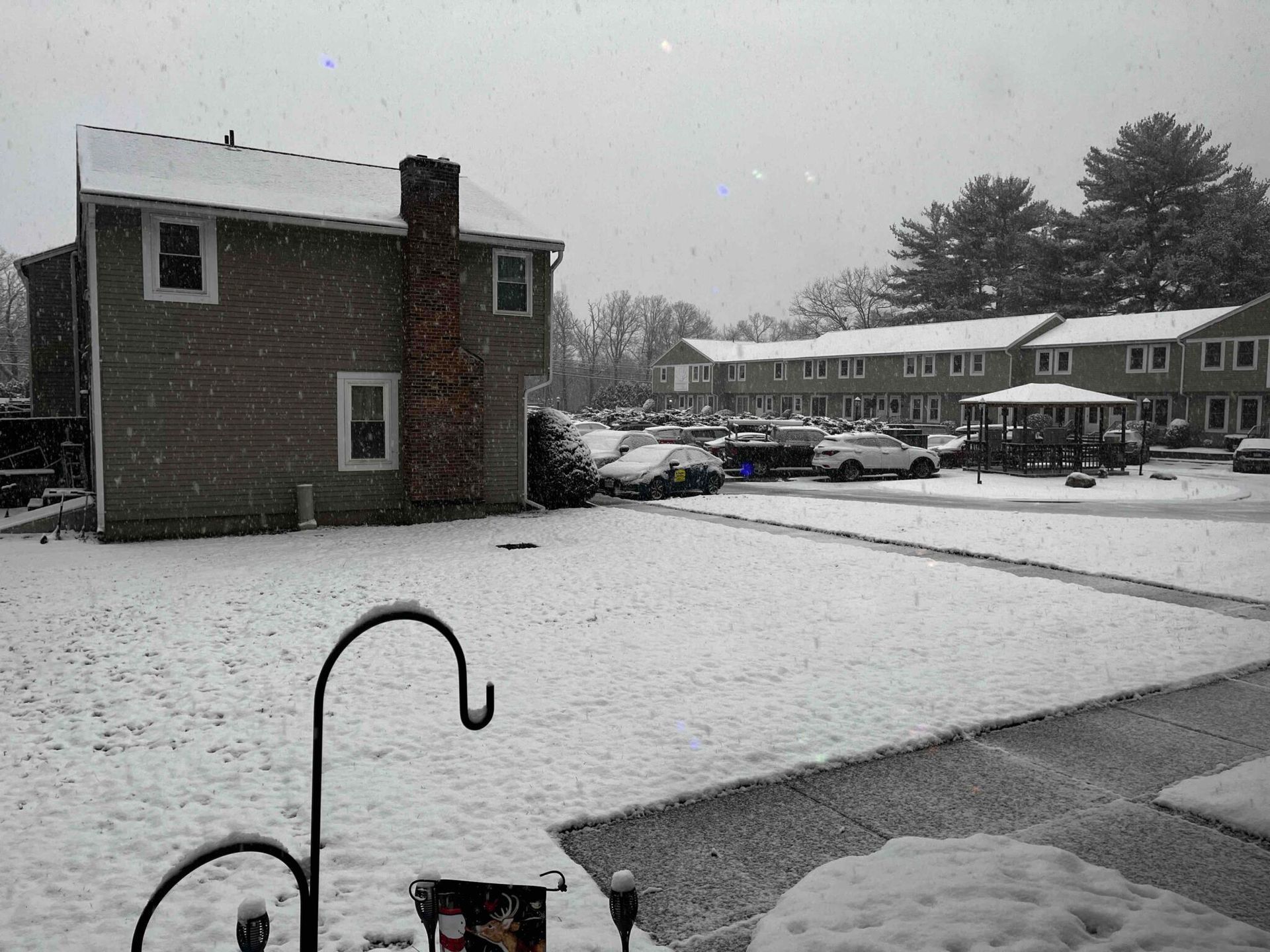 Snow-covered yard with houses in background, cars parked, and a gazebo. Snowflakes falling gently from sky.