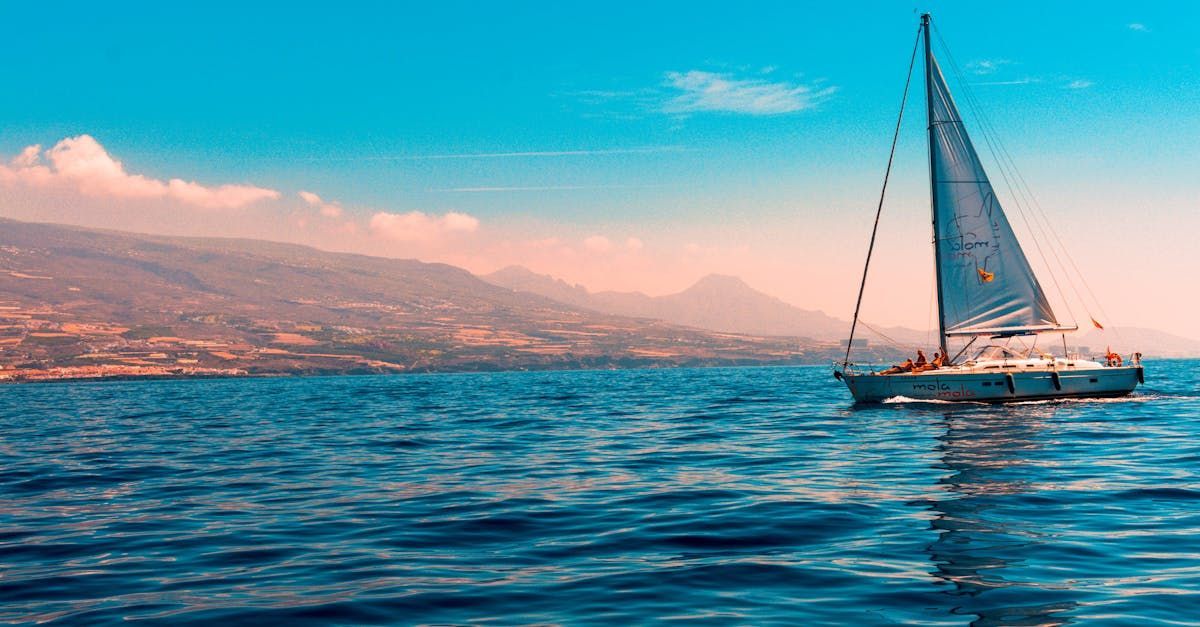 Sailboat gliding on blue sea with people aboard, mountains and clear sky in the background.