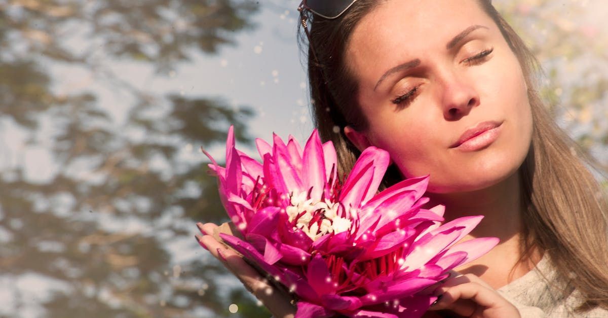 A female person holding vibrant pink flowers, with a blurred background of trees and sky reflecting in water.