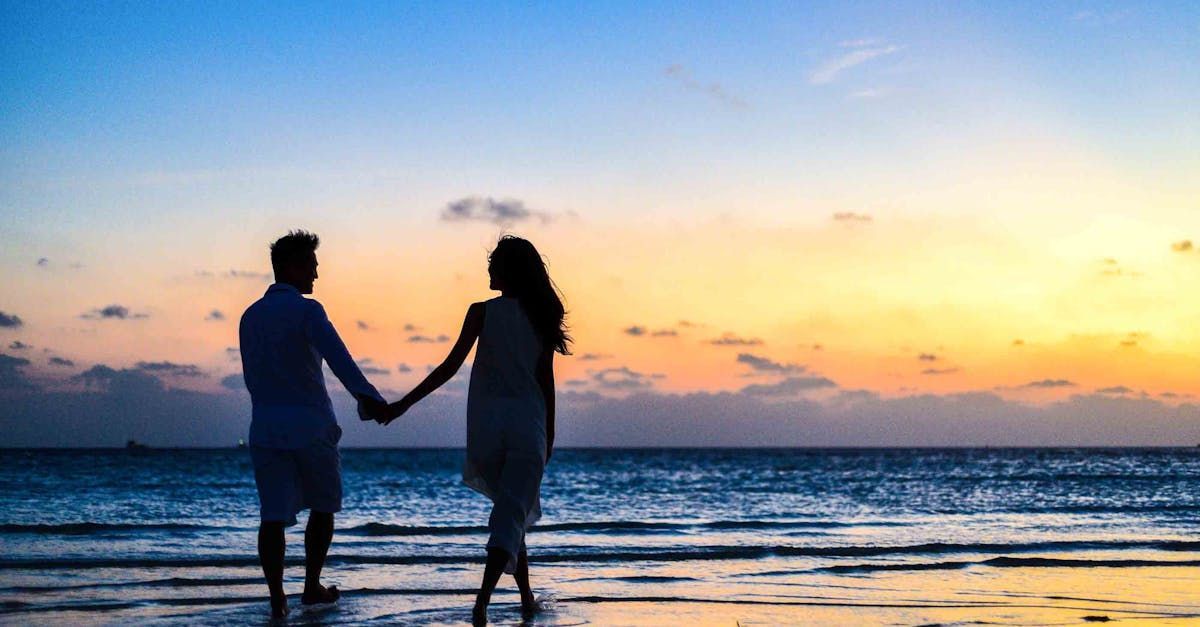 A couple holds hands while walking along the beach at sunset, silhouetted against a vibrant sky.