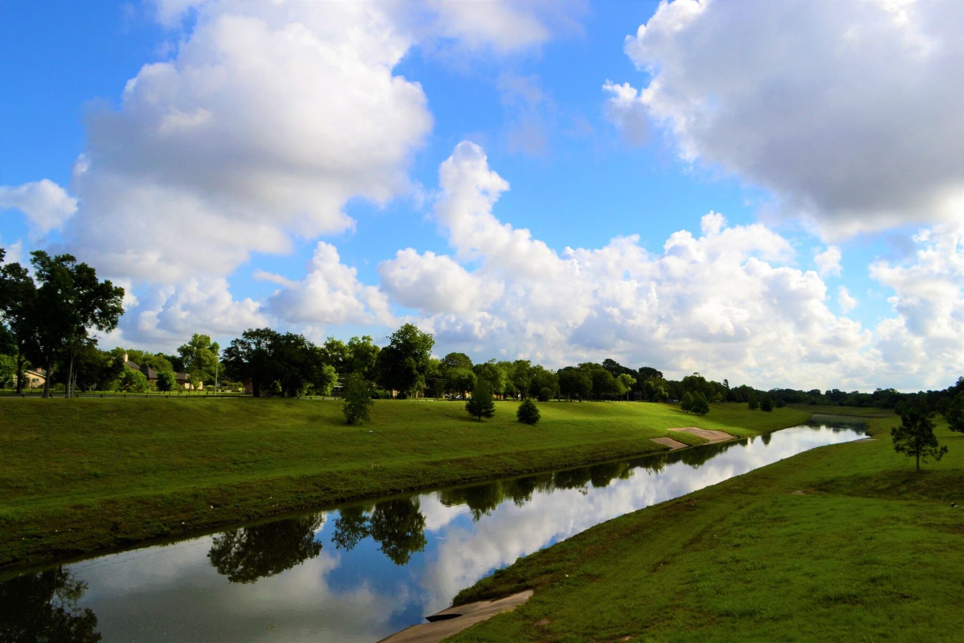 Lush green landscape with trees and a reflective river under a bright sky filled with fluffy white clouds.