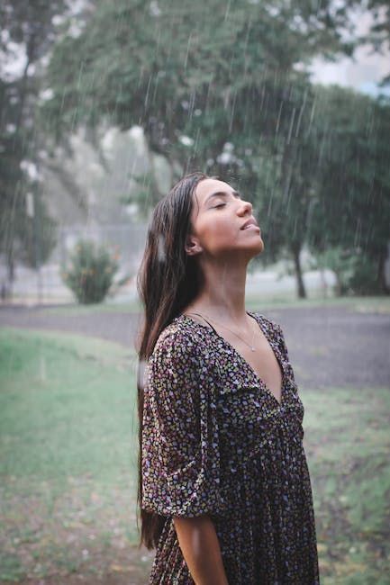 Woman in wet floral dress stands in the rain, facing upward, with long hair and trees blurred in the background.