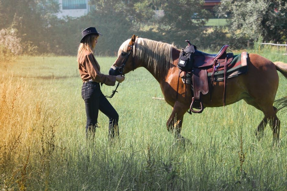 A Female person in a brown shirt and cowboy hat stands in a grassy field, holding the reins of a saddled brown horse.