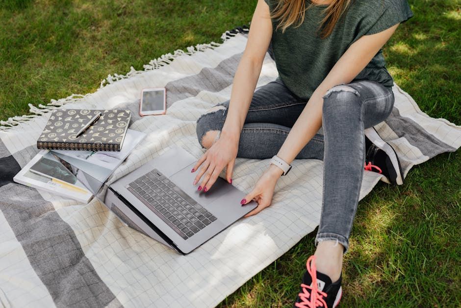 Woman sitting on a blanket in the grass, using a laptop, with a notebook, phone, and pens beside her.
