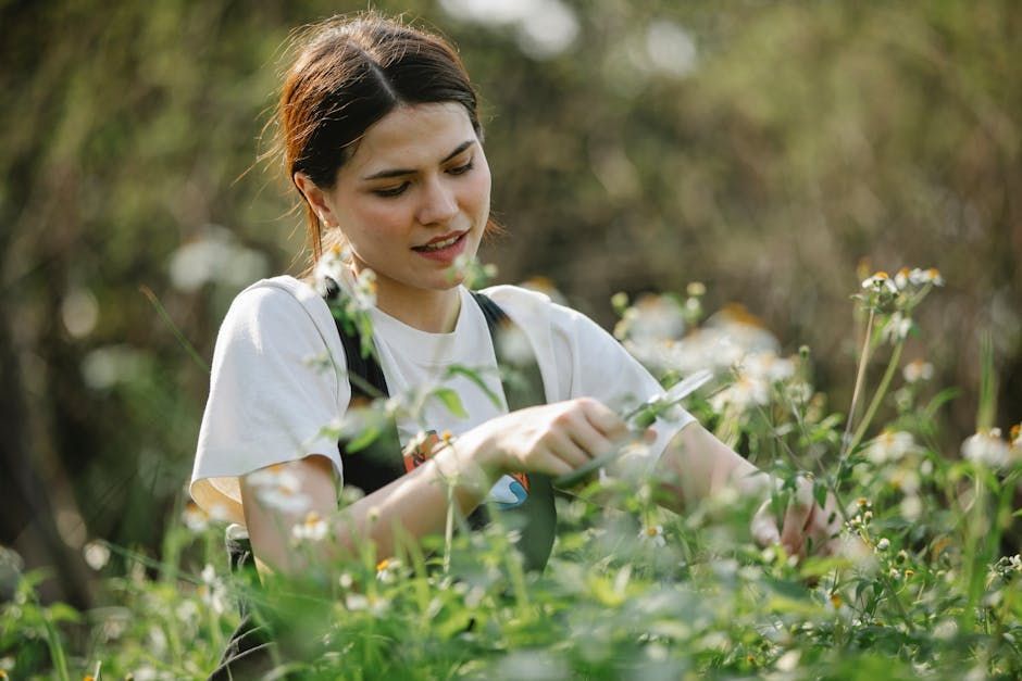 Female Person wearing a white shirt and green apron is gardening, surrounded by green plants and blurred natural background.