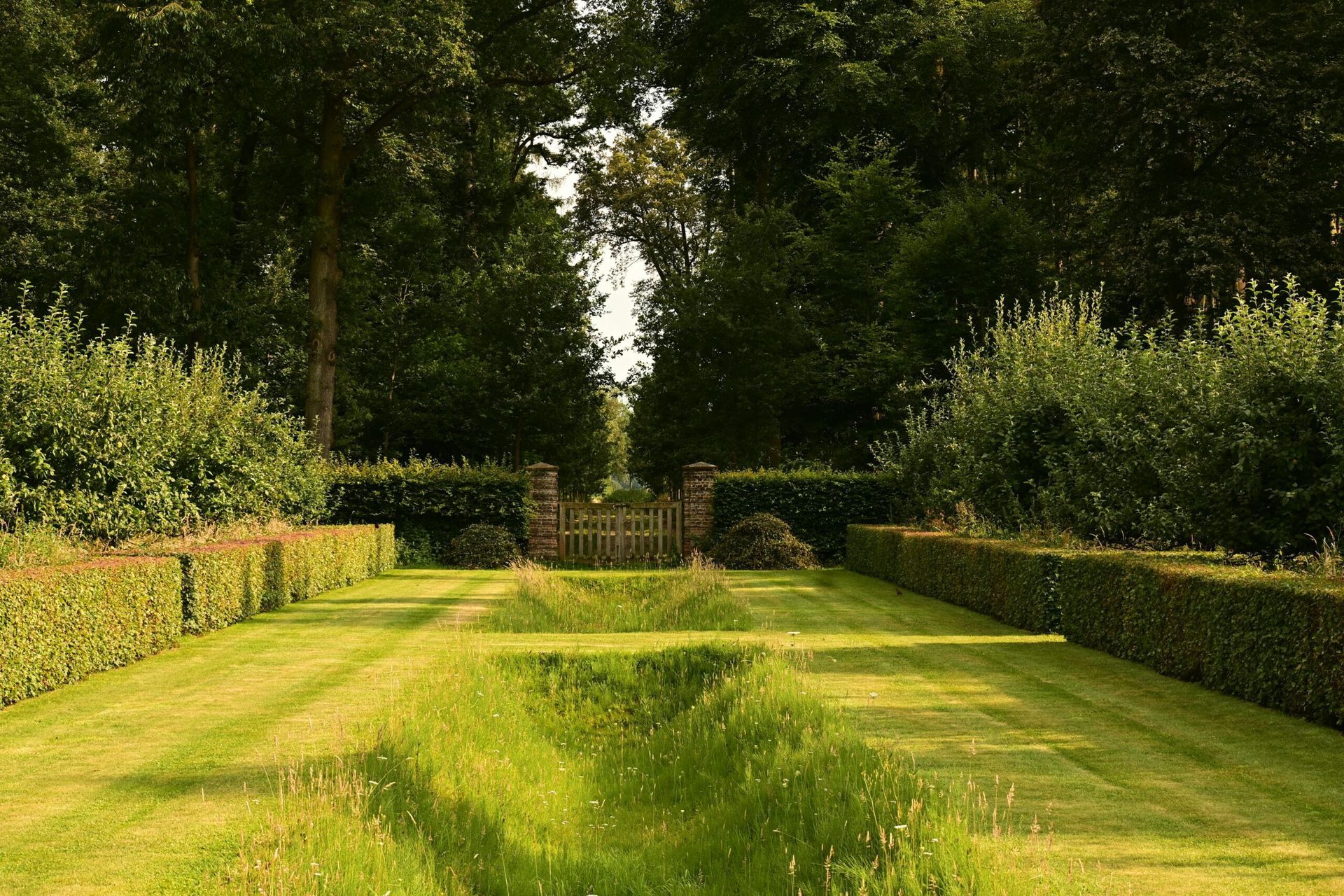 Manicured garden with trimmed hedges and lush grass, leading to a wooden gate flanked by tall trees in the background.
