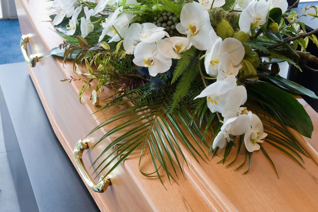 Arreglo floral funerario de orquídeas blancas sobre un ataúd de madera clara con asas doradas.