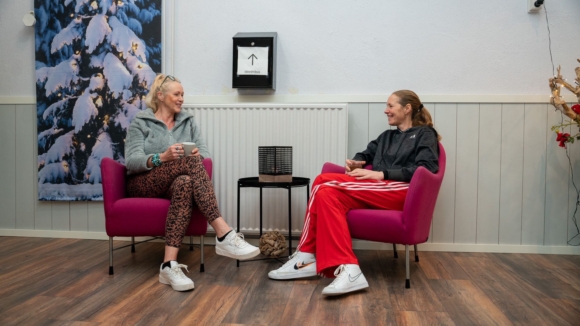Twee vrouwen zitten in fauteuils en praten. Interieur met een schilderij van een sneeuwlandschap.