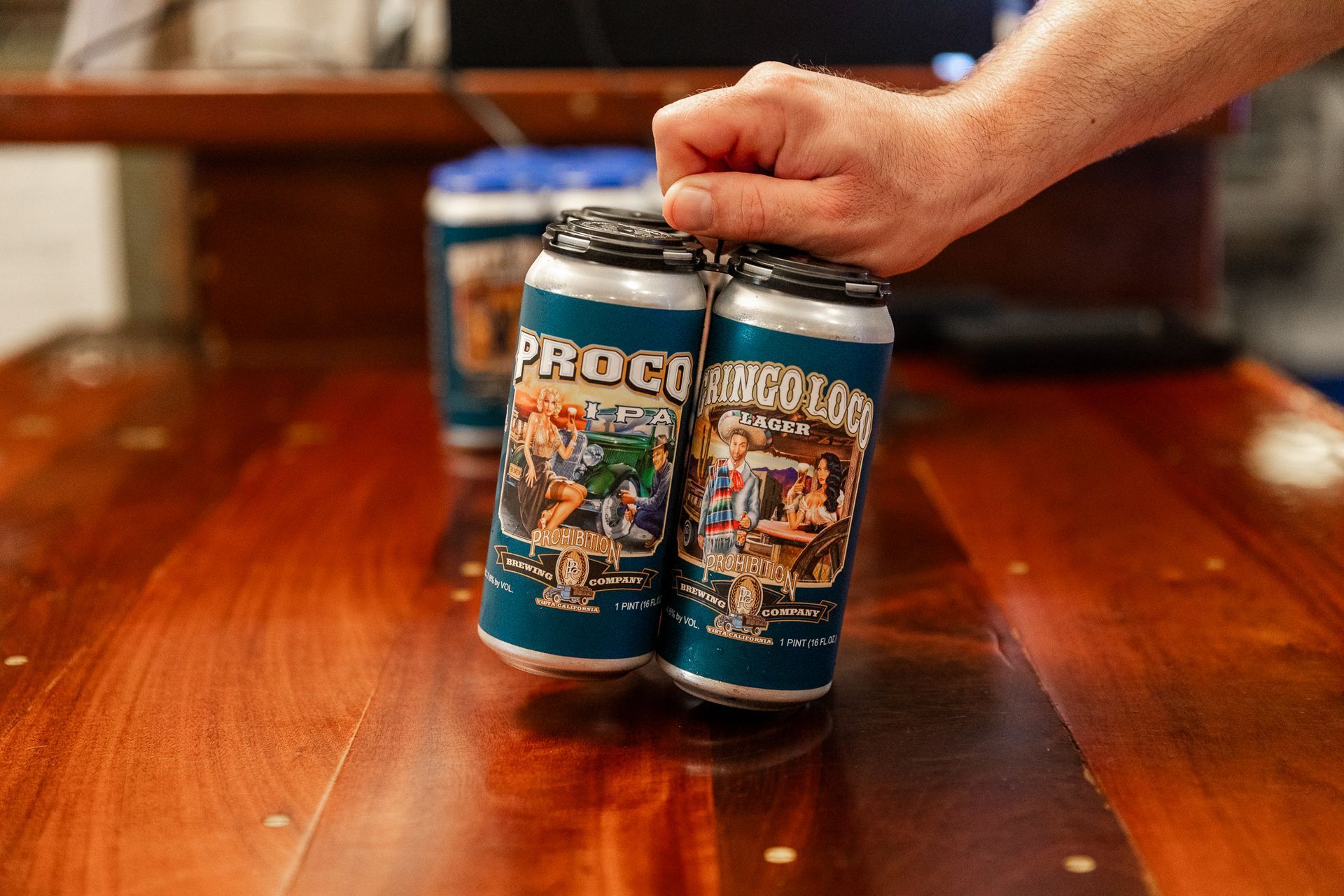 A person is holding four cans of beer on a wooden table.