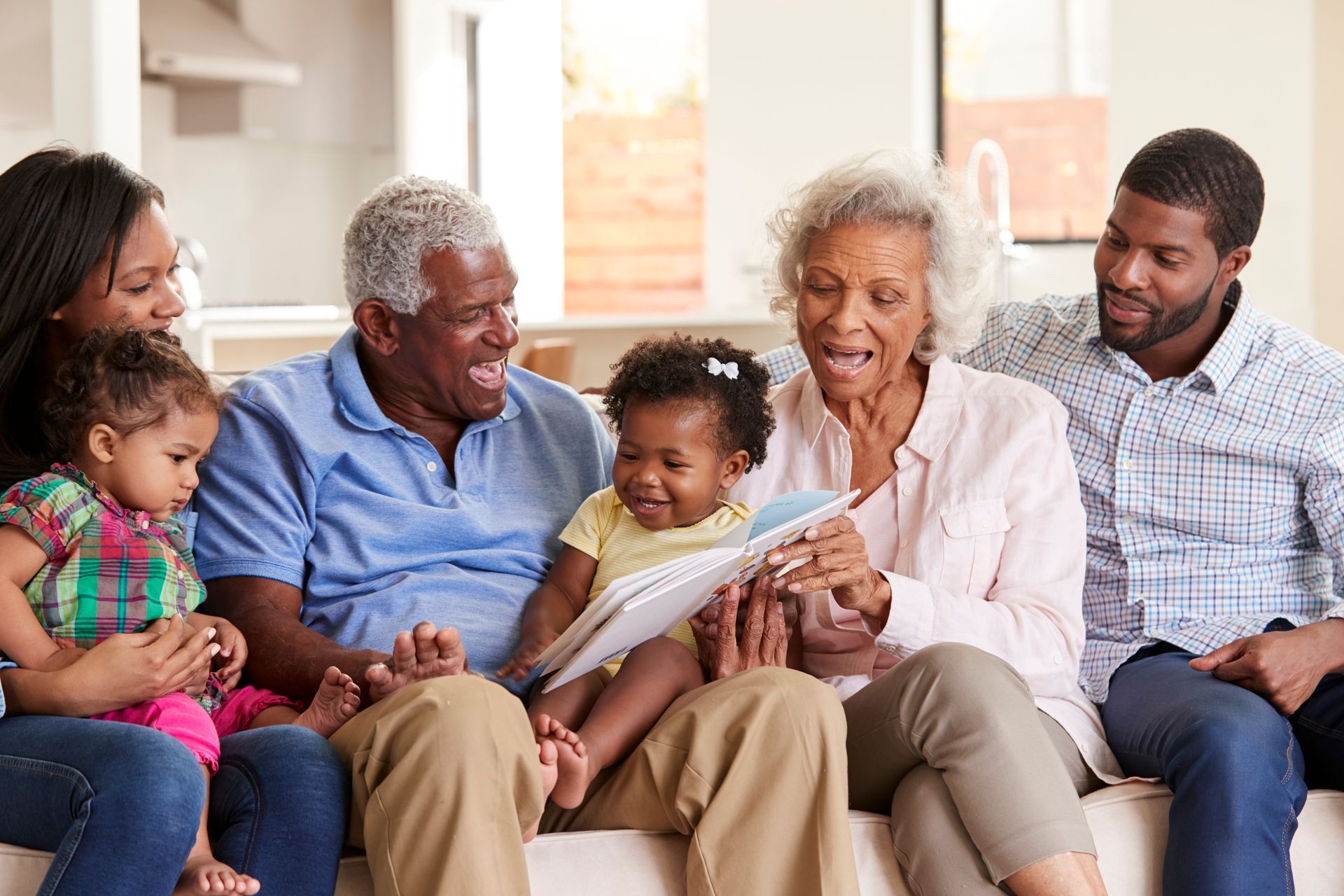 Family on a couch reading a book together; baby laughs, adults smile in a bright living room.