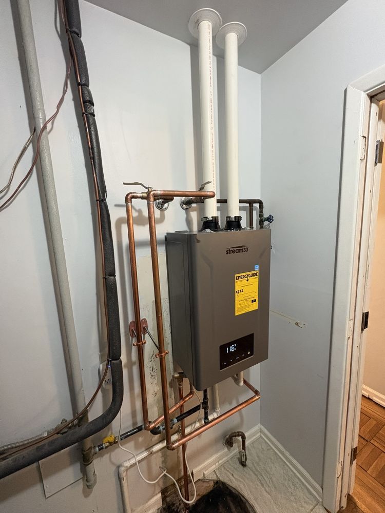 A wall-mounted, grey tankless water heater with copper pipes in a utility room. Two white exhaust pipes extend upwards.