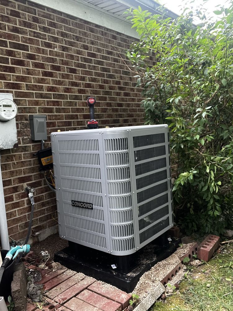 Gray air conditioning unit next to a brick wall and bushes.