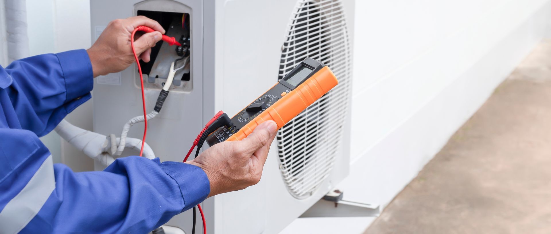 An electrician in blue overalls uses a multimeter to inspect an air conditioning unit.