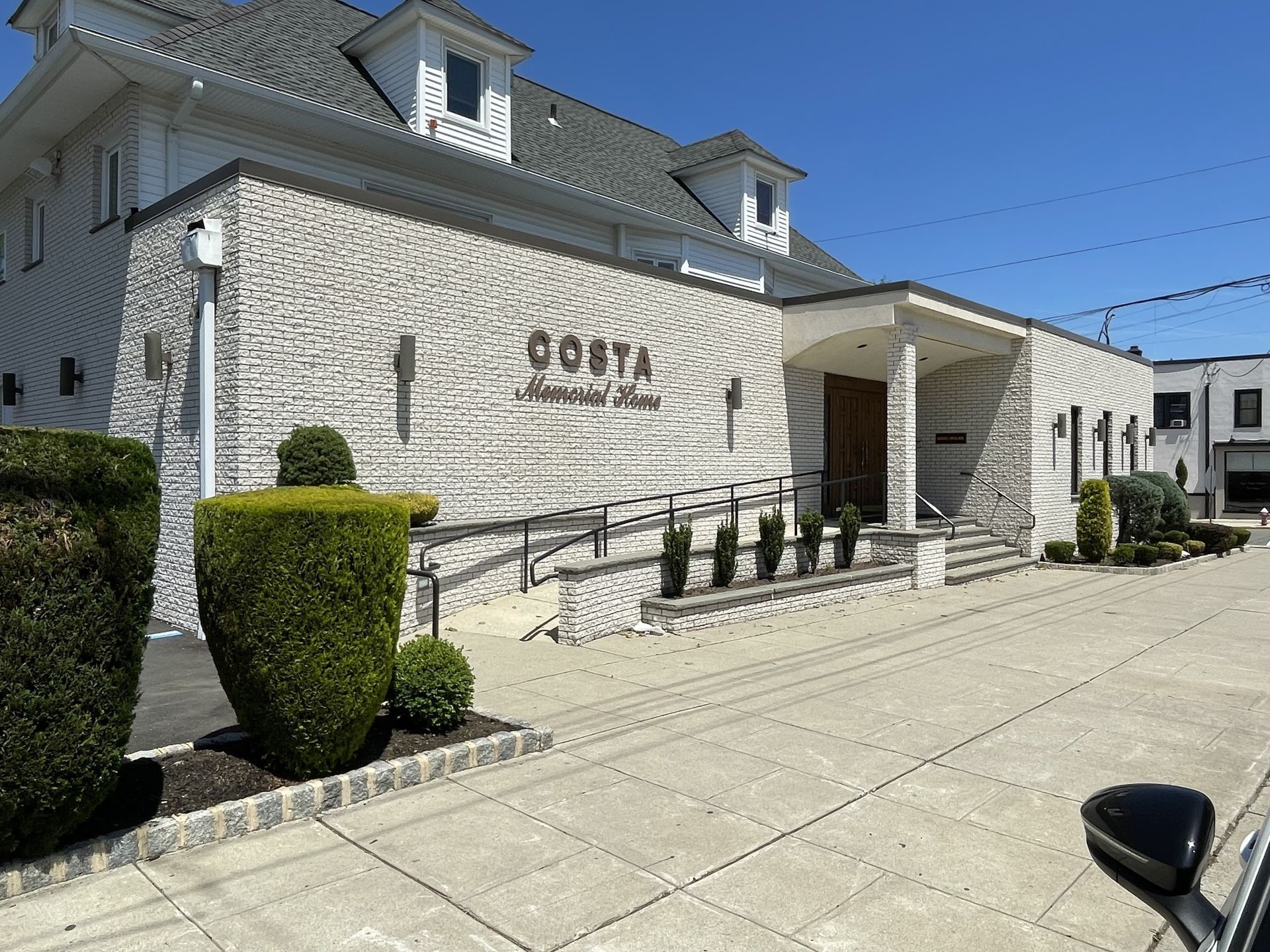Stone building with a small porch, trimmed hedges, and paved driveway under a clear blue sky