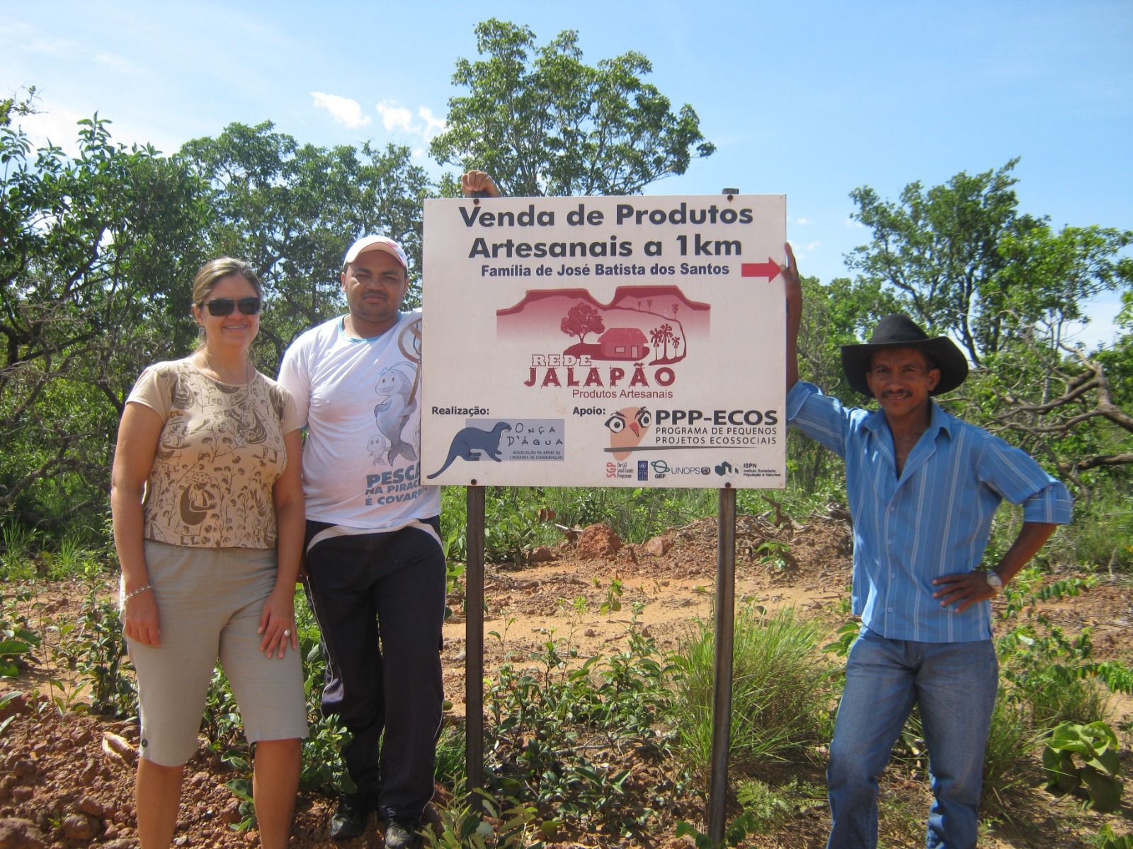 Três pessoas em pé em frente a uma placa que diz venda de produtos
