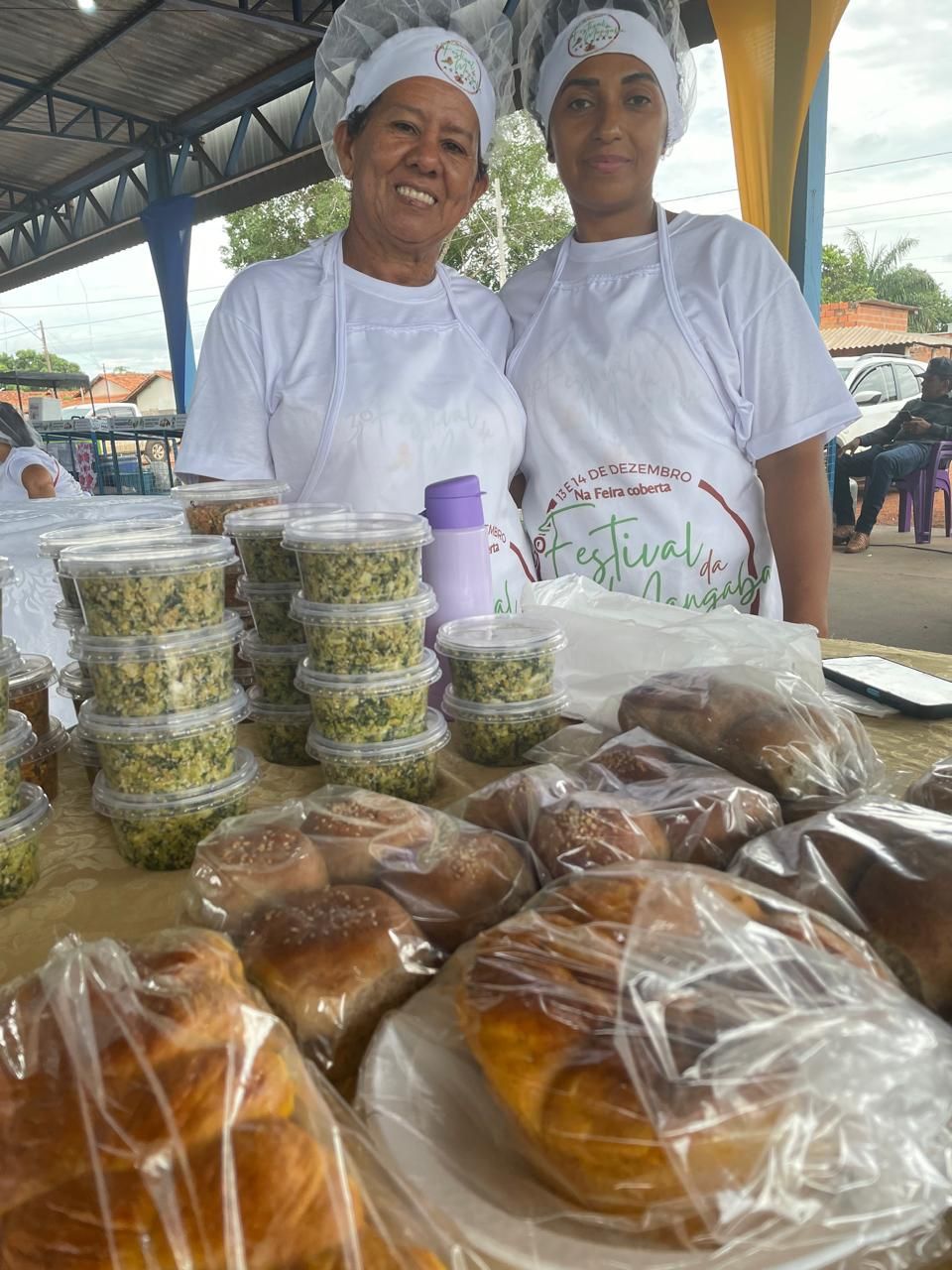 Duas mulheres estão de pé, uma ao lado da outra, em frente a uma mesa cheia de comida.