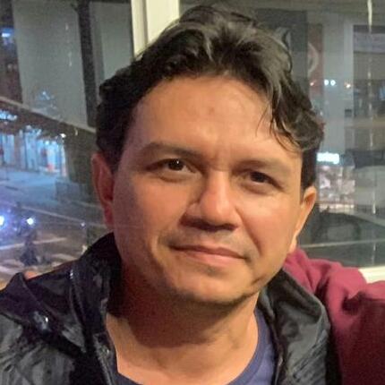Um homem vestindo uma jaqueta preta e uma camisa azul está sentado em frente a uma janela.