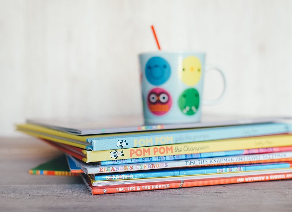 A stack of children 's books next to a mug that says pom pom