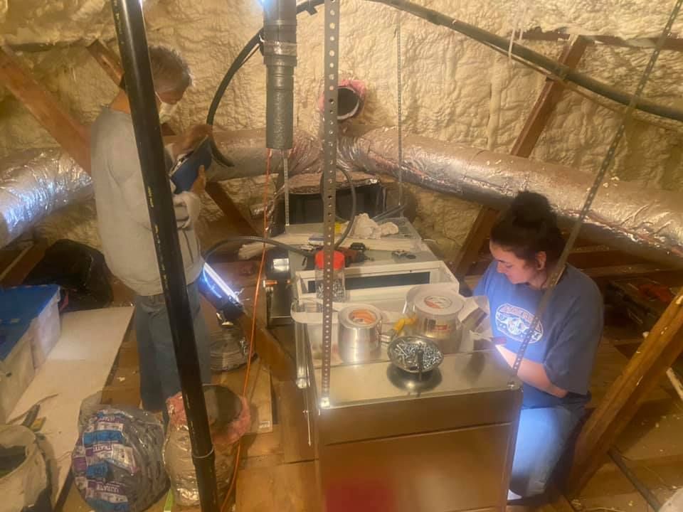A man and a woman are working on an air conditioner in a basement.