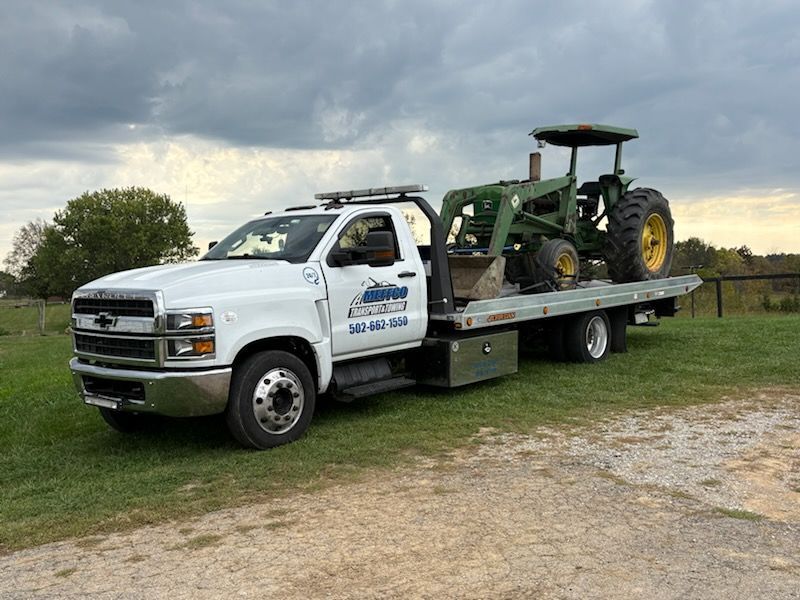 White tow truck transporting a green and yellow tractor on a rural grassy area. Overcast sky.