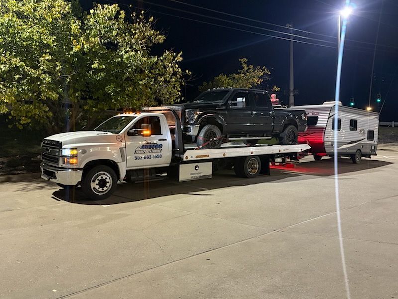 Tow truck hauling a black pickup truck and a travel trailer at night.