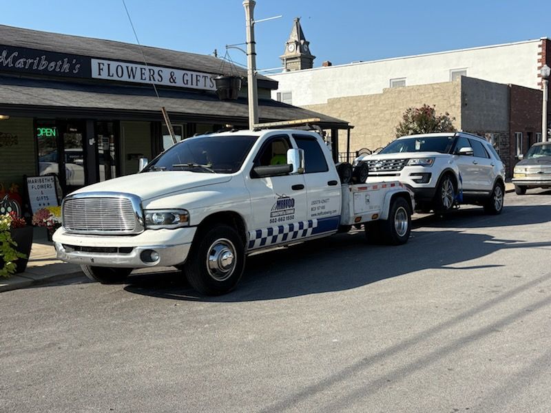 A white tow truck towing a white SUV on a sunny street, near a flower shop.