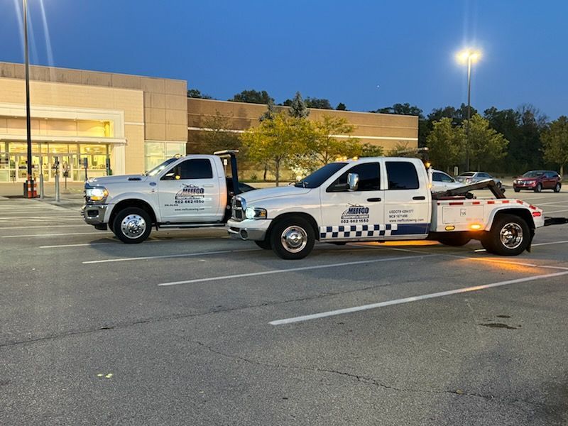 Two white tow trucks parked in a parking lot, one towing the other. A store is in the background.