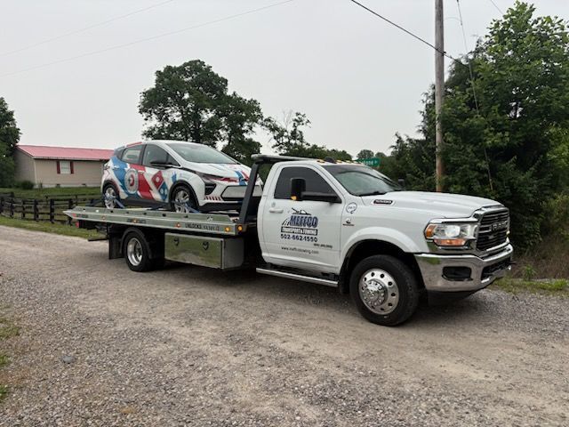 A white tow truck carries a white and decorated car on a gravel road, trees in the background.