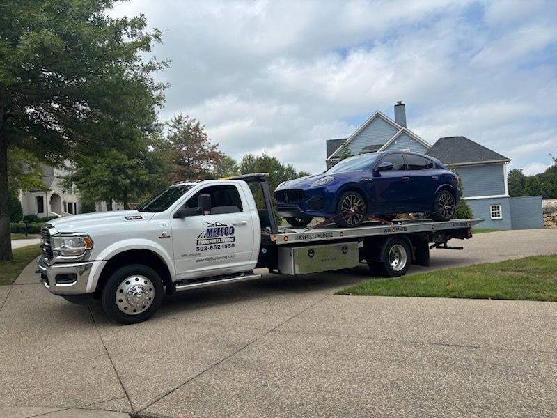 White tow truck carrying a blue car on a residential driveway on a cloudy day.