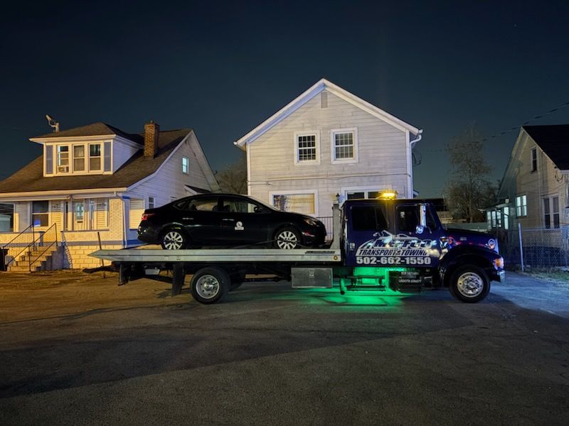 A black car loaded on a tow truck at night in front of houses; truck has green underglow.