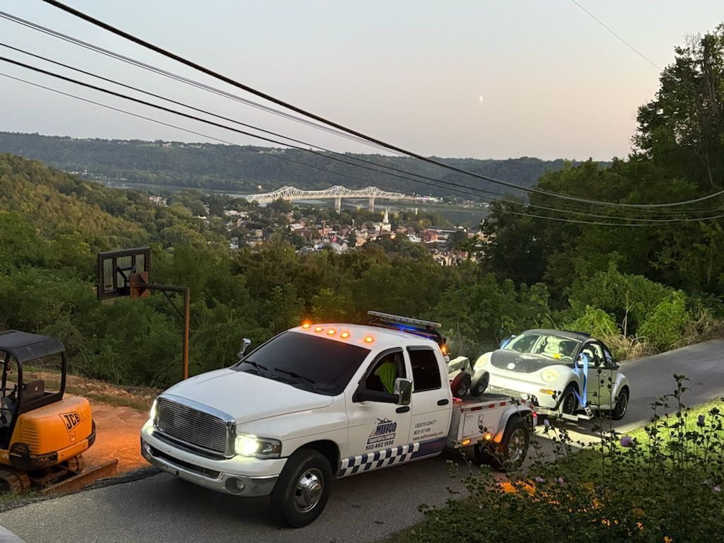 Tow truck towing a small car on a hillside road overlooking a town. Dusk, overhead power lines.