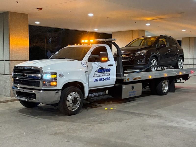 White tow truck hauling a black SUV, parked under a building with flashing amber lights.