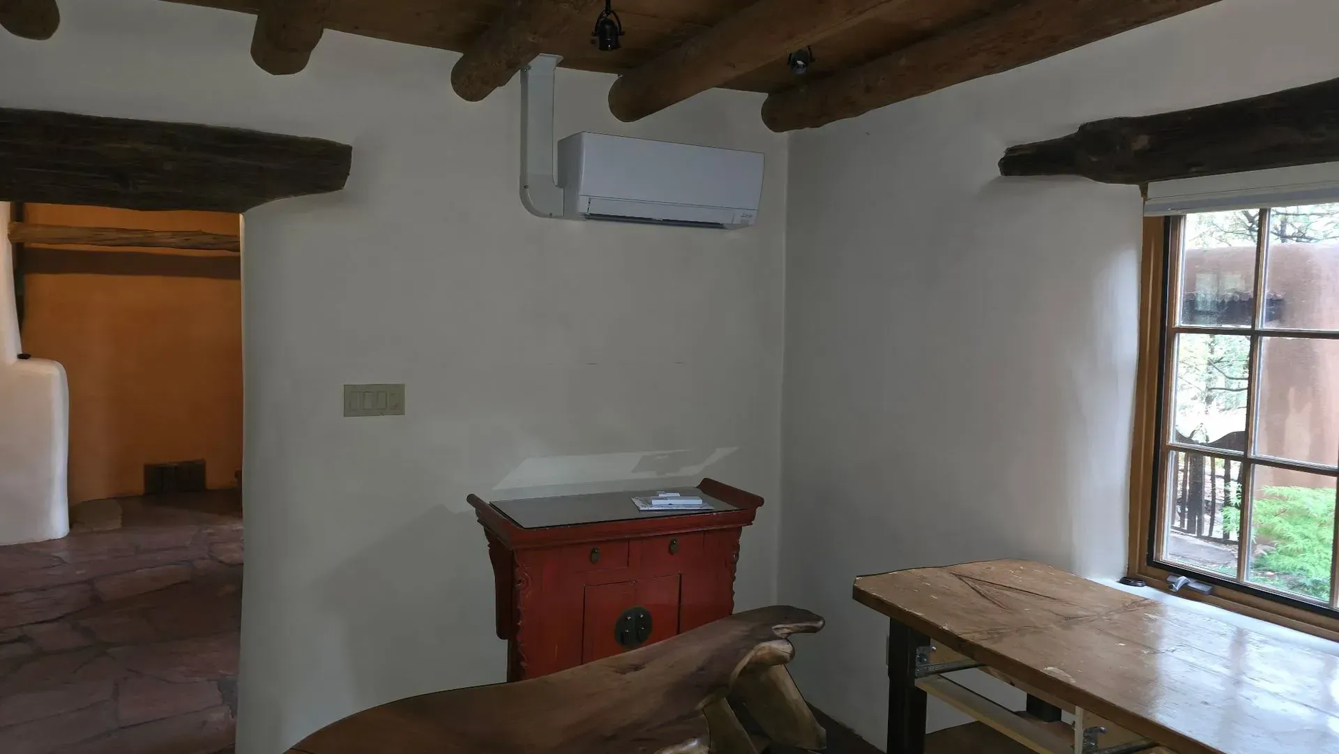Interior corner of a room with white walls, wooden beams, a red cabinet, and a window overlooking greenery.