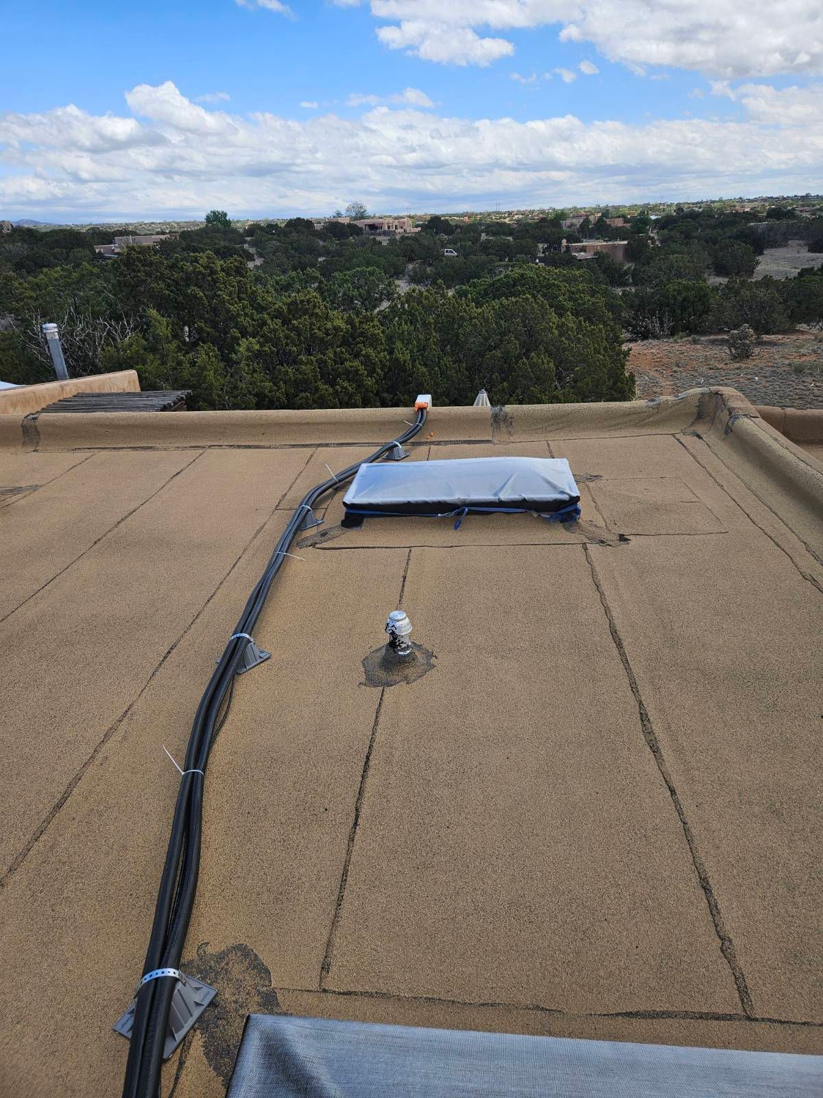 Rooftop view with black cables, a covered skylight, and trees in the distance under a partly cloudy sky.