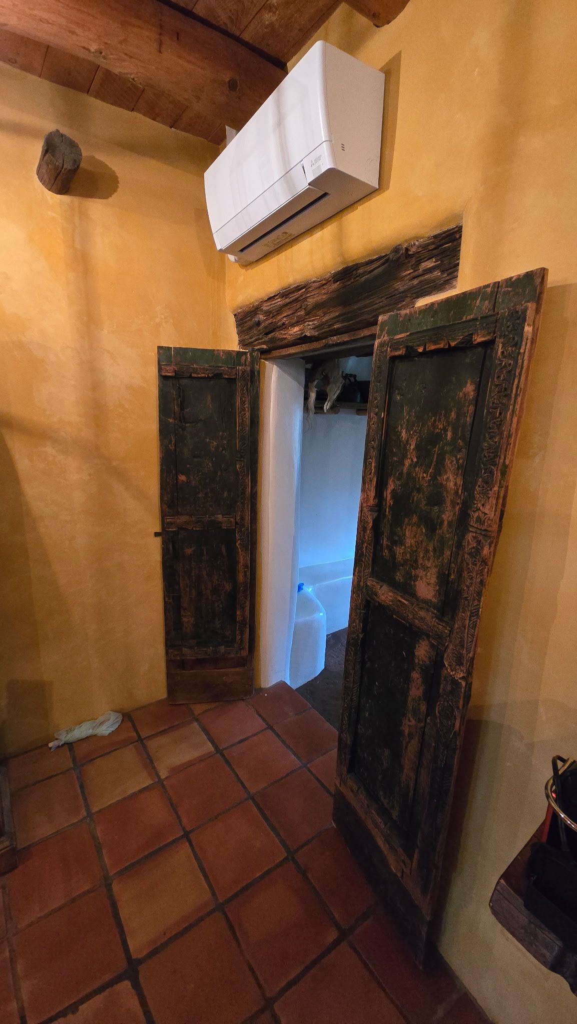 Rustic doorway with worn, dark wooden doors. Opens into a white room. Orange tiled floor and yellow walls.