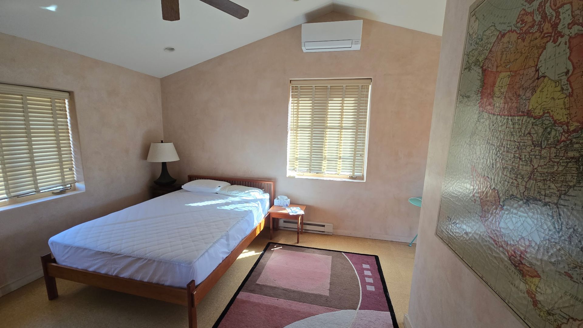 Bedroom with a single bed, small rug, and window with blinds. Pink walls, a lamp, and map art.