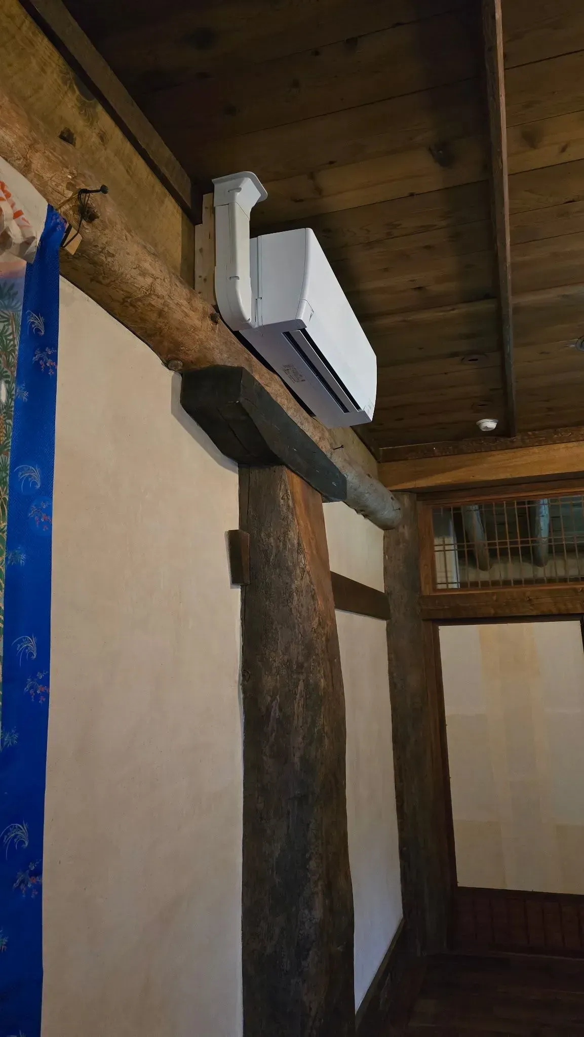 White air conditioner mounted on wooden beam inside a building with visible wooden ceiling and column.