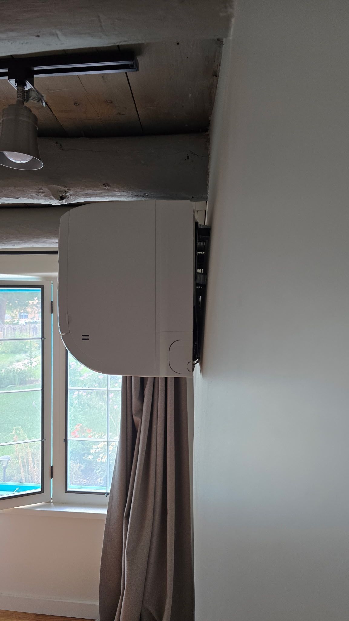 White air conditioning unit mounted on a white wall in front of a window with curtains.
