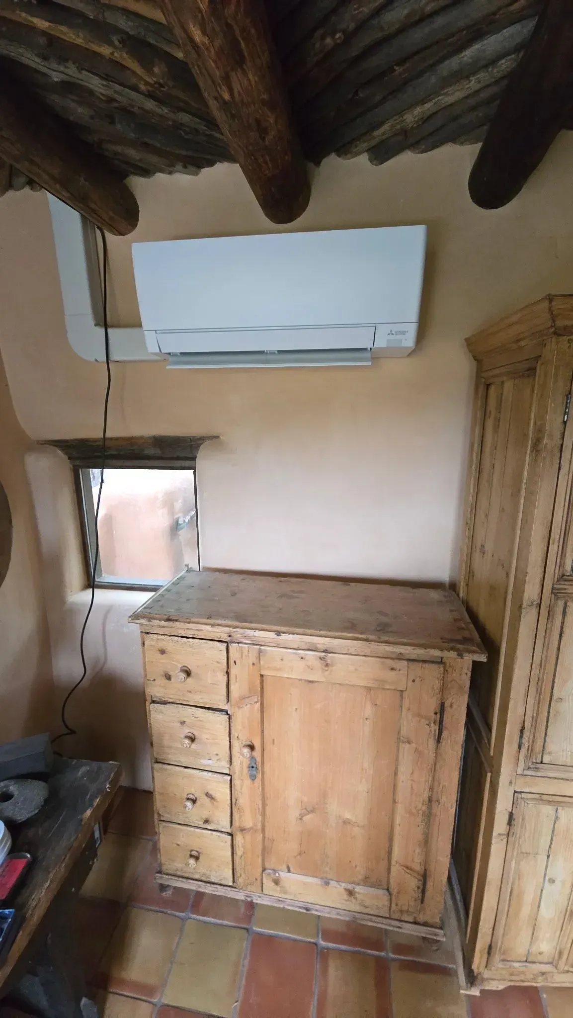 Air conditioning unit above wooden cabinet in rustic room; exposed ceiling beams.