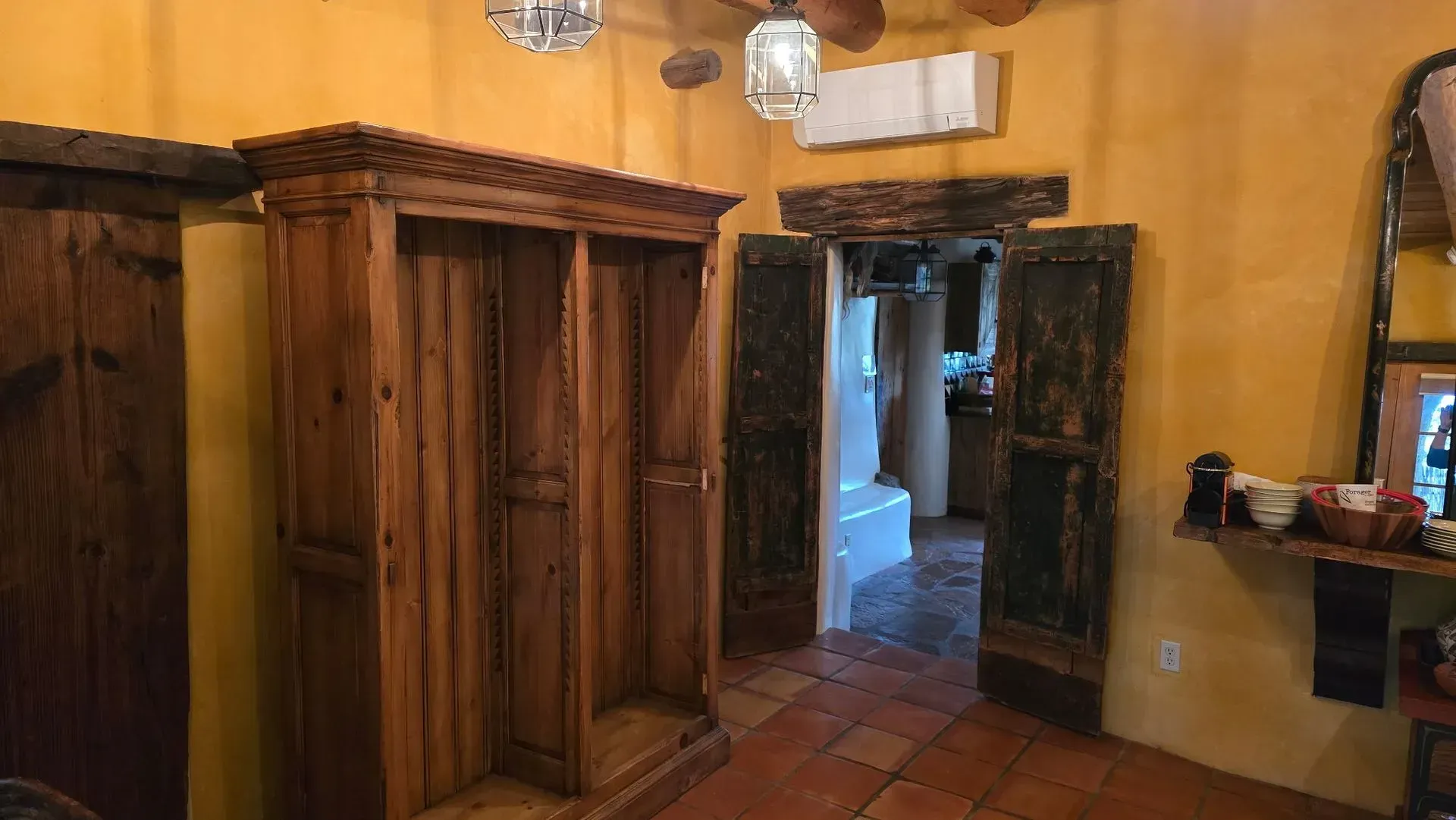 Wooden armoire and doorway in a room with terracotta tile flooring and yellow walls.