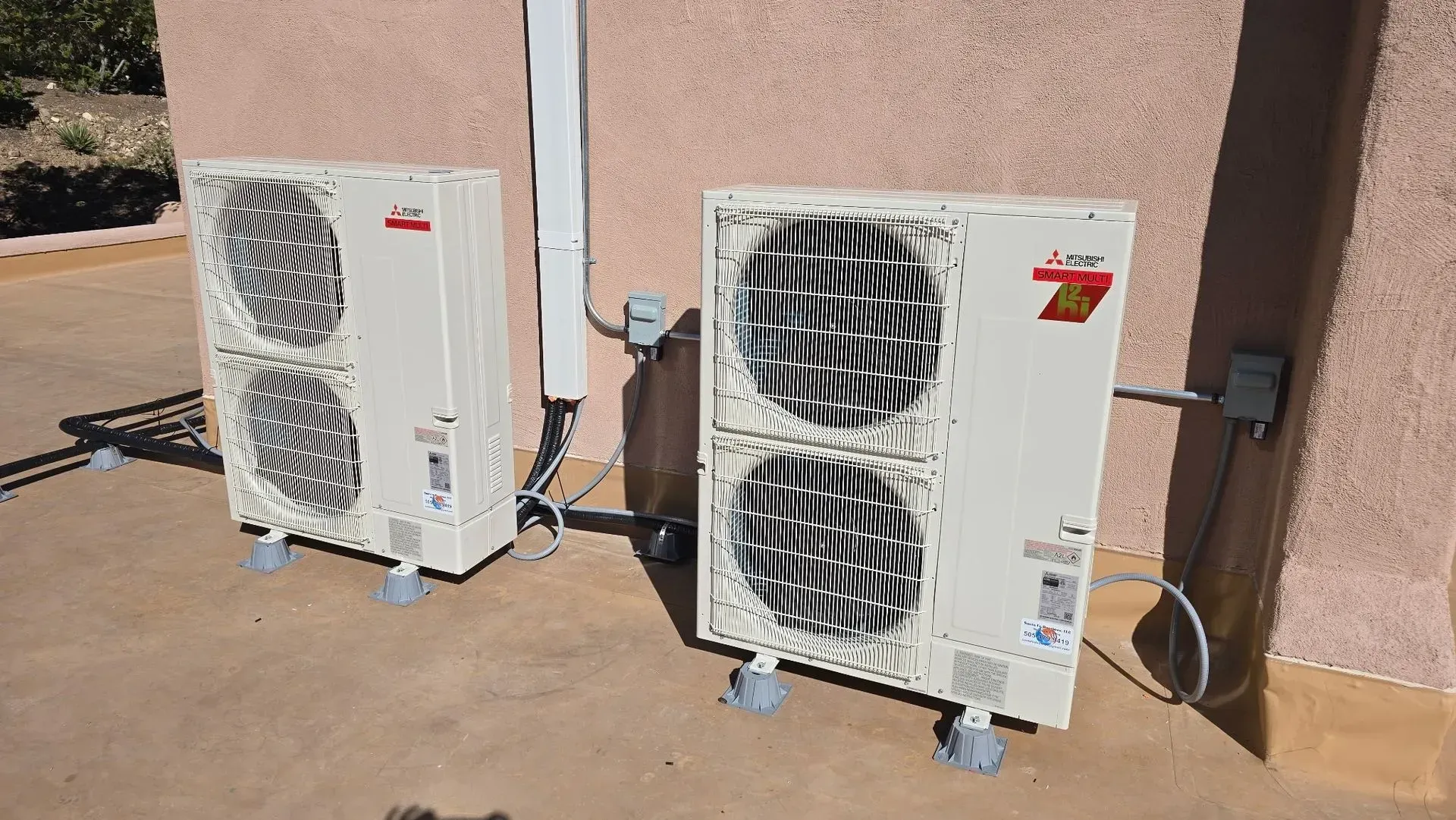 Two beige air conditioning units outside, on a concrete surface, against a light brown wall.