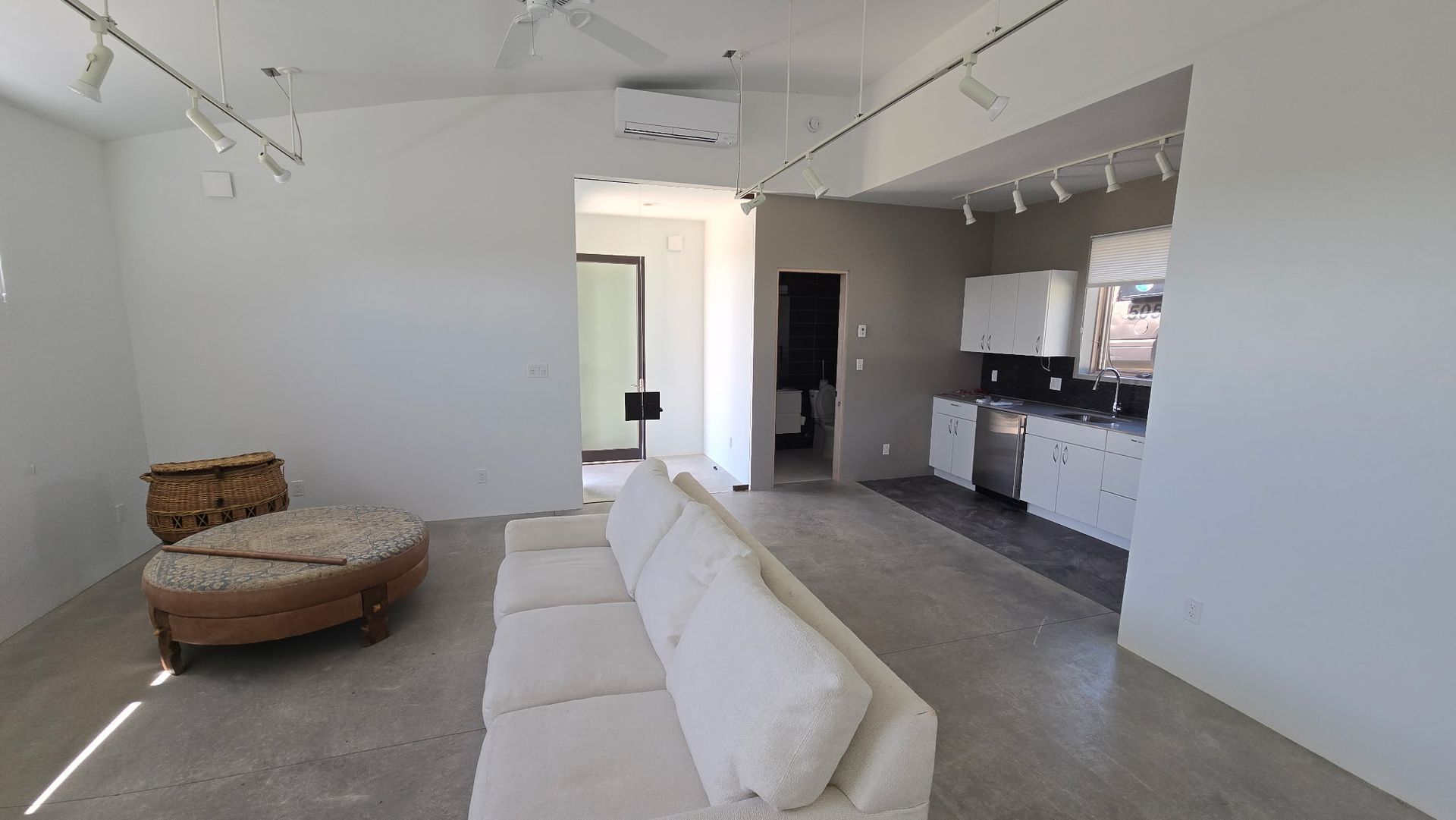Spacious living room with white sofa, kitchen with white cabinets, and an open doorway to another room.