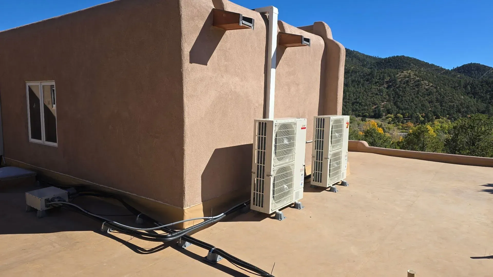 Two air conditioning units mounted on a flat, brown roof of a stucco building; mountains in background.