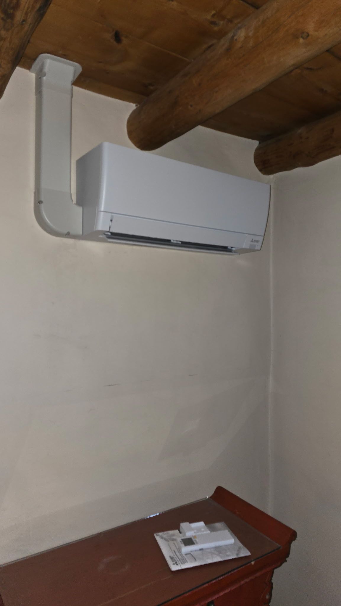 White air conditioner unit mounted on a beige wall, next to a wood beam ceiling. A brown cabinet sits below.