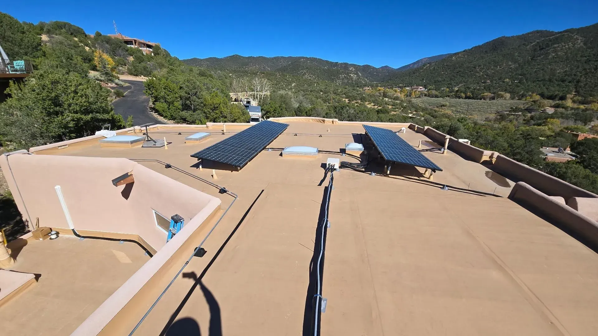 Solar panels on a flat, tan roof with a mountainous, forested backdrop under a blue sky.